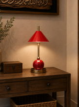 Load image into Gallery viewer, A red glass table lamp with a red conical shade and brass base sits on a dark wood console table in a warmly lit interior setting.