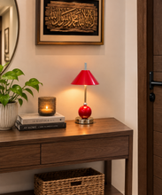 Load image into Gallery viewer, A red table lamp with a conical shade and glossy red base sits on a dark wood console table in a warmly lit entryway. A potted plant, stacked books, a candle, and framed wall art are visible around it.