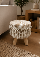 Load image into Gallery viewer, A round woven ottoman stool with a thick cream rope body, decorative fringe around the lower half, and four light wood legs, shown in a styled living room next to a sofa and wooden side table.