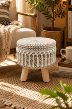 Load image into Gallery viewer, A round woven pouf stool with a cream and white patterned body and light wood legs, styled in a cozy sunlit living room next to a chair and bookshelf.