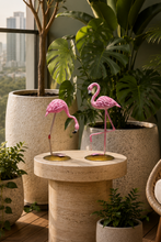 Load image into Gallery viewer, A styled outdoor patio scene with two pink flamingo figurines standing on a round stone side table, surrounded by potted plants, a wicker chair, and a balcony view.