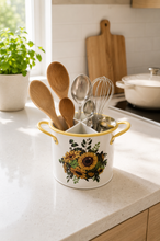 Load image into Gallery viewer, A white ceramic kitchen utensil holder with yellow handles and a sunflower floral print, filled with wooden and metal kitchen tools, sitting on a light speckled countertop in a bright kitchen.