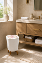 Load image into Gallery viewer, A white ribbed cylindrical trash can with a floral lid design and wooden legs, styled in a bright, modern bathroom next to a wooden vanity.