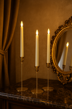 Load image into Gallery viewer, Three lit candles in gold candle holders on a reflective surface with a mirror and curtain in the background.