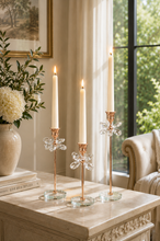 Load image into Gallery viewer, Crystal Bloom 3-piece rose gold candle holder set with lit taper candles on marble side table by window