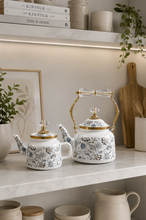 Load image into Gallery viewer, Floral enamel tea kettle in white with blue-grey floral pattern, gold-tone detailing and crystal-style accents, available in 1-liter and 2.4-liter capacities.