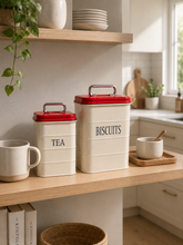 Load image into Gallery viewer, Retro Pantry metal Tea and Biscuit canister set with cream bodies, glossy red lids and silver-toned carrying handles on kitchen shelf – MetaphorBD