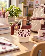 Load image into Gallery viewer, Cocoa Cream leopard-print water bottle with burgundy handle, cream carrying strap and flower charm on a study desk.