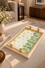 Load image into Gallery viewer, Botanical tray with ivory daisies, green leaves, gold-tone framing and transparent handles on a neutral upholstered ottoman.