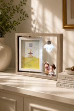 Load image into Gallery viewer, A light wood shadow box frame displayed on a cream-colored cabinet. The frame contains a small colorful illustration of a carousel and two miniature figurines, with a small hanging bulb casting a soft shadow.
