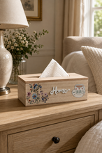 Load image into Gallery viewer, A rectangular wooden tissue box holder with a floral design, the word 'Home' in script, and a teacup graphic, holding white tissue paper on a light wood bedside table in a cozy bedroom setting.

