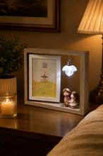Load image into Gallery viewer, A rustic wooden shadow box night light sits on a wooden bedside table, illuminated by a warm candle and a bedside lamp. The shadow box features a small framed illustration of a carousel and a hanging light bulb that glows with a cool blue-white light, accompanied by two tiny doll figures.
