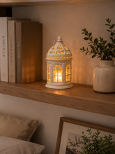 Load image into Gallery viewer, A white decorative lantern with a warm yellow light glowing inside, styled on a wooden shelf next to books and a potted plant.
