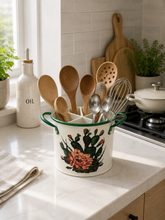 Load image into Gallery viewer, A white enamel kitchen utensil holder with green handles and a floral cactus print, filled with wooden and metal kitchen utensils, sitting on a white marble kitchen countertop.
