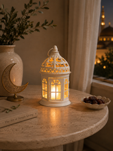 Load image into Gallery viewer, A white ornate metal lantern with a warm yellow glow inside, styled on a round stone table next to a vase, a gold crescent moon sculpture, and a bowl of dates.
