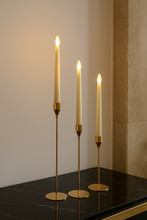 Load image into Gallery viewer, Three lit candles in gold candle holders on a dark surface with a neutral background
