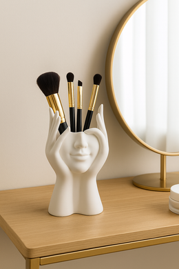 Face-shaped white ceramic makeup brush holder on vanity with gold-trimmed brushes

