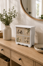 Load image into Gallery viewer, White and wood-tone 4-drawer decorative mini organizer cabinet with botanical, bicycle and LOVE designs on dressing table.
