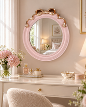 Load image into Gallery viewer, Pink 18 × 18 inch round wall mirror with sculpted bow and gold-tone accents above a dressing table.
