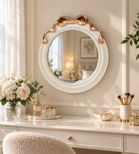 Load image into Gallery viewer, White 18 × 18 inch round wall mirror with sculpted bow and gold-tone accents above a dressing table.
