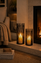 Load image into Gallery viewer, Two black metal wicker-style candle holders with lit candles, placed on a marble fireplace hearth in a cozy living room setting.
